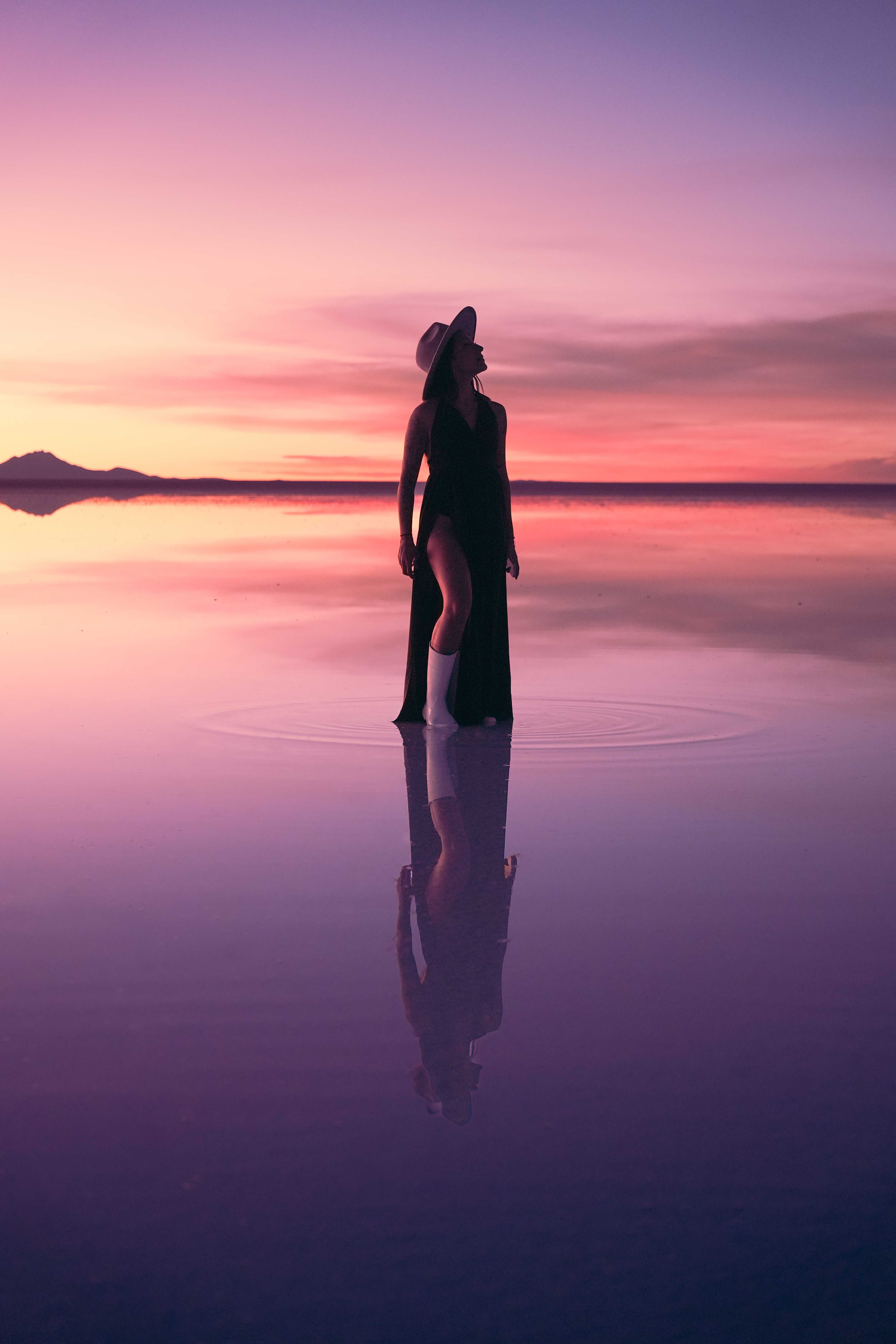 Silueta de una mujer con sombrero sobre el Salar de Uyuni al atardecer, reflejada en el agua.